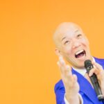青いスーツ姿で笑顔のまま手を差し出す鴨頭嘉人