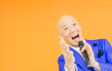 青いスーツ姿で笑顔のまま手を差し出す鴨頭嘉人