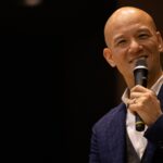 暗いステージでマイクを持ち、笑顔で話す鴨頭嘉人