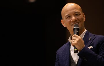 暗いステージでマイクを持ち、笑顔で話す鴨頭嘉人