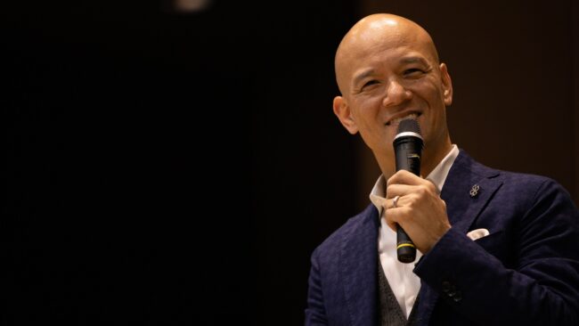 暗いステージでマイクを持ち、笑顔で話す鴨頭嘉人