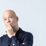 白い背景で腕を組み、口元に手を添えて考える鴨頭嘉人