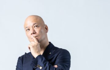 白い背景で腕を組み、口元に手を添えて考える鴨頭嘉人