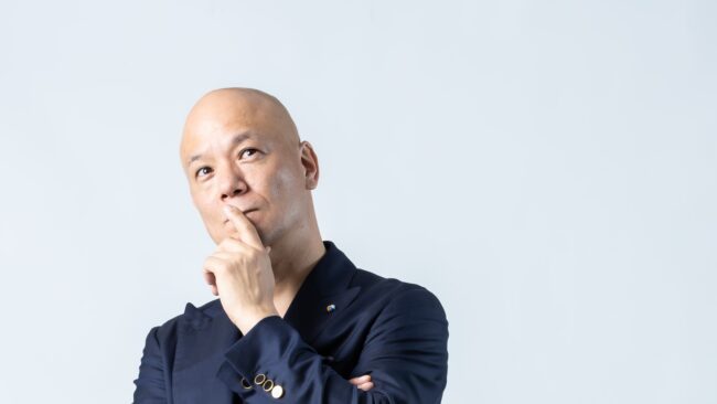 白い背景で腕を組み、口元に手を添えて考える鴨頭嘉人