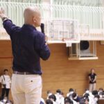 学校の体育館で大勢の学生へ向けて話す鴨頭嘉人