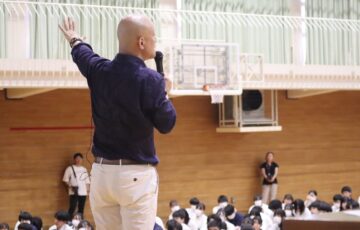 学校の体育館で大勢の学生へ向けて話す鴨頭嘉人