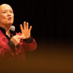 ステージで赤いジャケットを着て力強く話す鴨頭嘉人