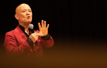 ステージで赤いジャケットを着て力強く話す鴨頭嘉人