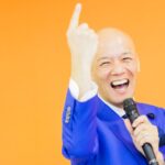 青いスーツ姿で笑顔のまま指を上げて話す鴨頭嘉人