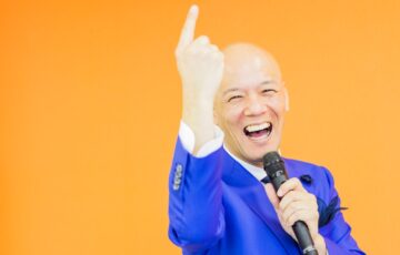 青いスーツ姿で笑顔のまま指を上げて話す鴨頭嘉人