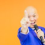 青いスーツ姿で笑顔のまま手を差し出す鴨頭嘉人