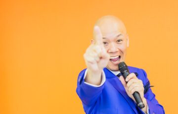 青いスーツ姿で笑顔のまま手を差し出す鴨頭嘉人