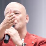 赤いジャケット姿で聴衆へ手を伸ばしながら話す鴨頭嘉人