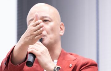 赤いジャケット姿で聴衆へ手を伸ばしながら話す鴨頭嘉人